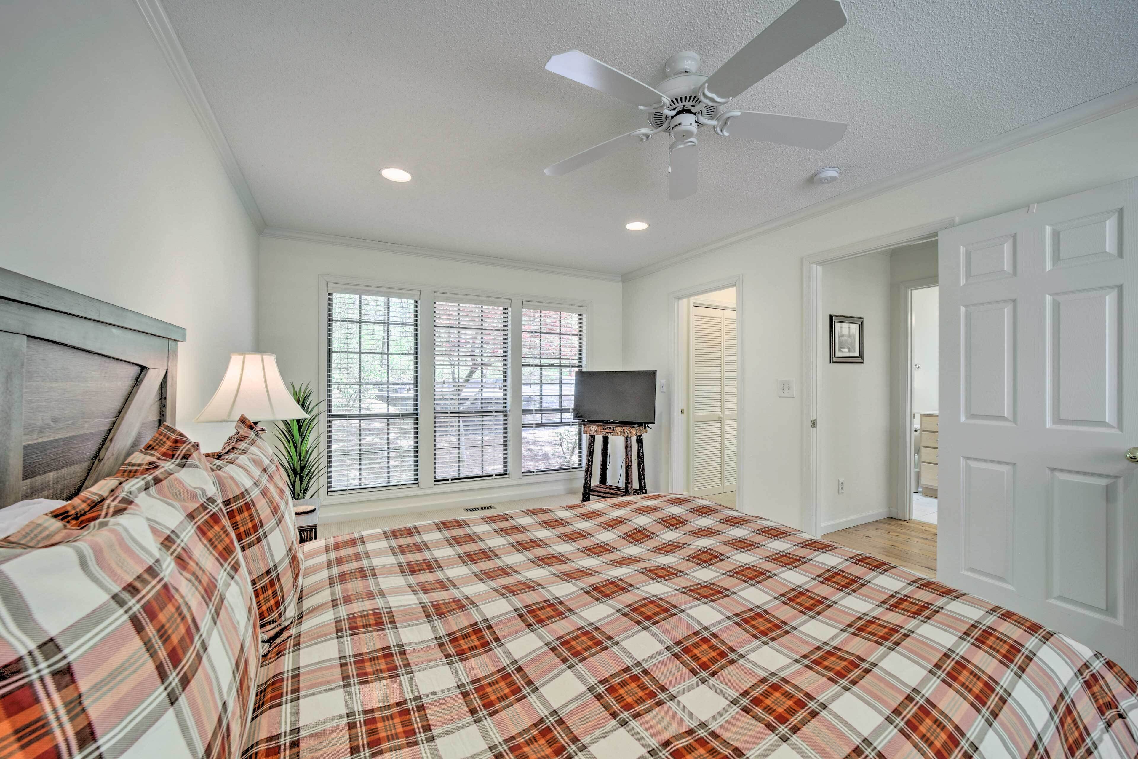 A bedroom with a plaid bedspread, a ceiling fan, a TV on a stand, and a window with blinds.