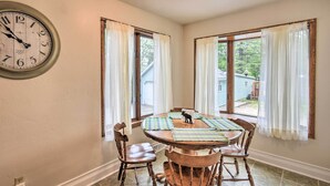A dining room with a round wooden table, four wooden chairs, a clock on the wall, and two windows with curtains. - Walkable Lake Elkhart Apt: Dine, Shop, Swim! (Elkhart Lake)