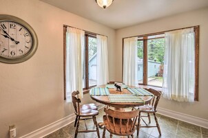 Comedor con una mesa redonda de madera, cuatro sillas de madera, un reloj en la pared y dos ventanas con cortinas.