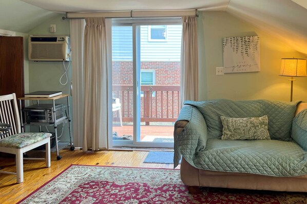 A cozy living room with a green sofa, a wooden bench, a red and white patterned rug, and a sliding glass door leading to a backyard.