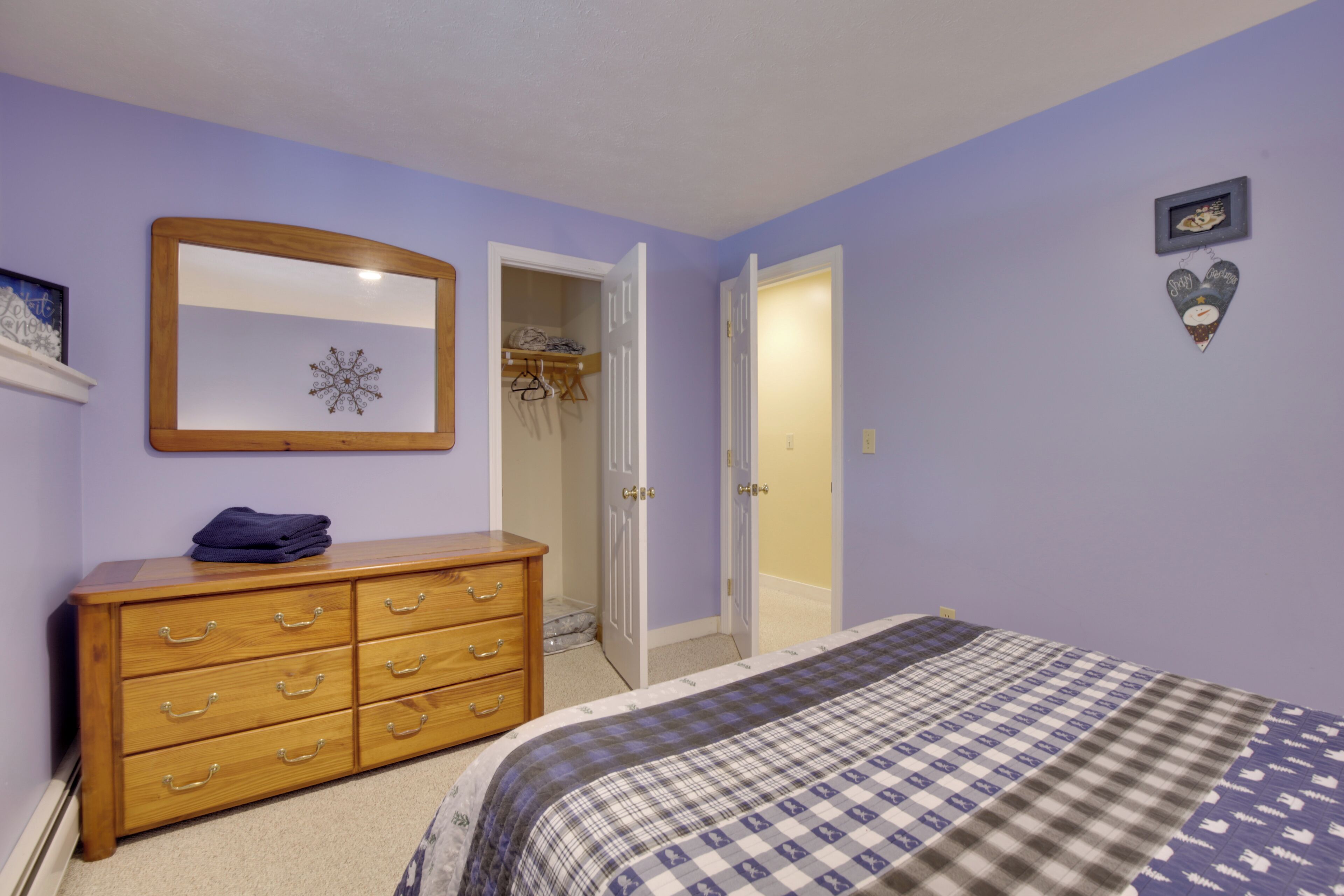 Une chambre à coucher avec une commode en bois, un lit recouvert d’une couverture à carreaux, un miroir et une armoire où sont suspendus des vêtements.