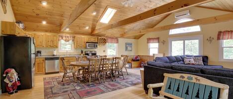 A spacious living area with a wooden ceiling, a dining table with chairs, a sofa, and a bench with a forest-themed design.