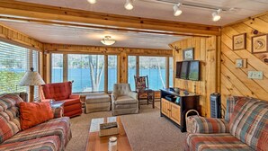 L’intérieur chaleureux d’un chalet avec des murs en bois, un téléviseur, une cheminée et une grande fenêtre donnant sur un lac.