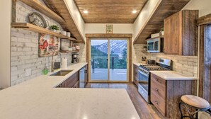 Une cuisine avec des meubles en bois, un grand îlot central et une vue sur les montagnes, visible à travers une porte vitrée.
