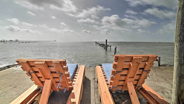 Two wooden lounge chairs with blue cushions on a concrete deck overlooking a body of water with a pier in the distance. - Rockport Bayfront Studio w/ Patio: 'casa Familia' (Rockport)