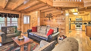 A cozy log cabin interior with a living room featuring a leather sofa, a green armchair, and a wooden coffee table. A kitchen with wooden cabinets and a microwave is visible in the background.