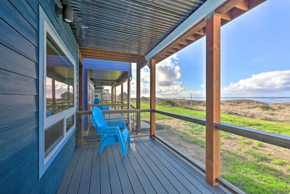A wooden deck with blue chairs, overlooking a beach with dunes and a cloudy sky.