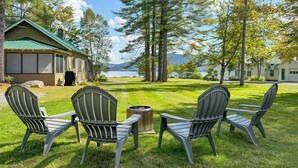 A grassy outdoor area with four Adirondack chairs facing a metal fire pit, with a cabin and a lake in the background. - Lake Pleasant Cabin on 3 Acres w/ Private Beach! (Speculator)