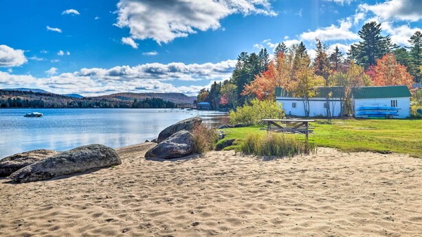 A lakeside scene with a sandy beach, a wooden bench, and a small cabin with a green roof. - Lake Pleasant Cabin on 3 Acres w/ Private Beach! (Speculator)