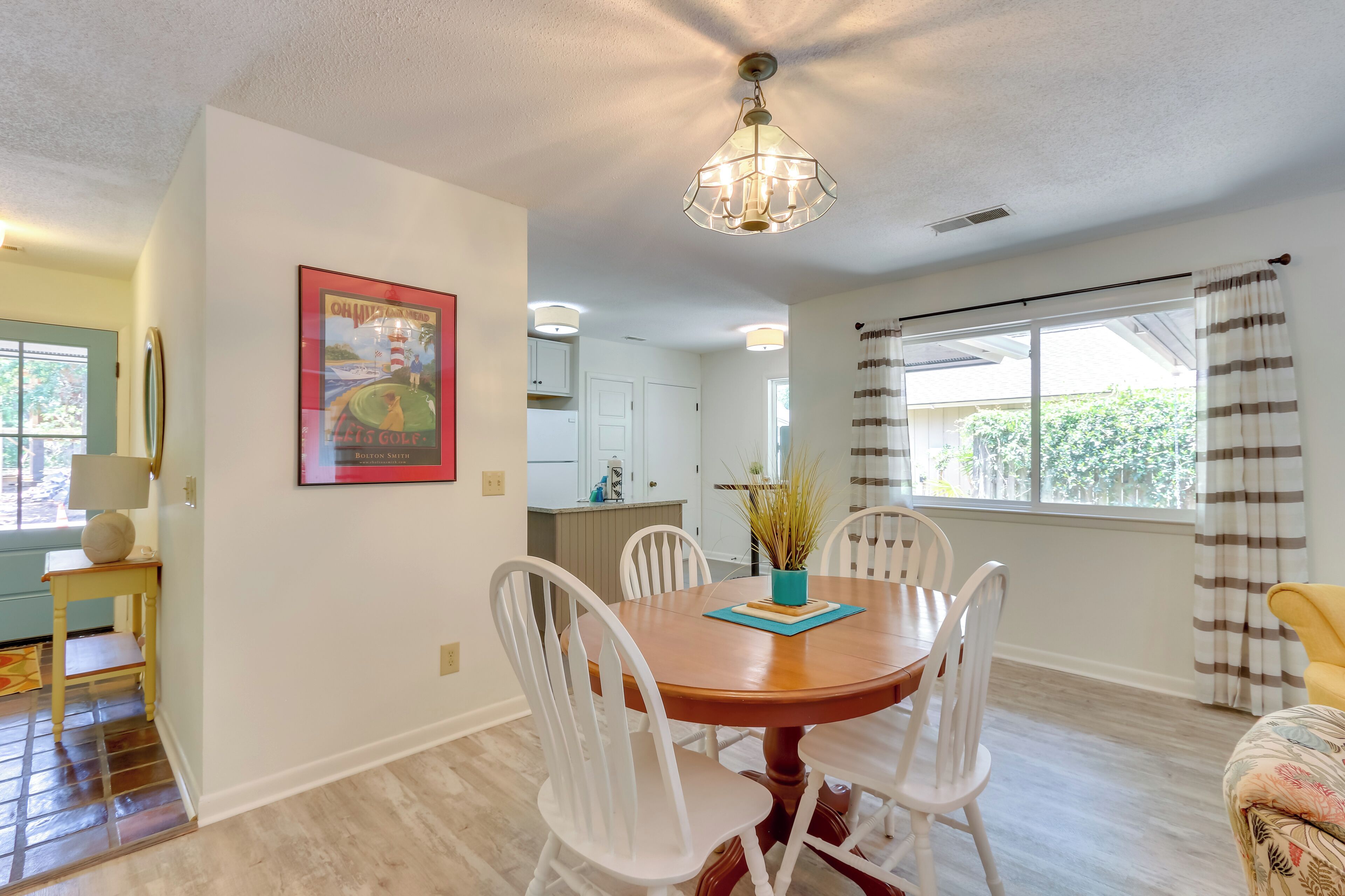 A dining area with a wooden table and white chairs, a chandelier, and a kitchen in the background.