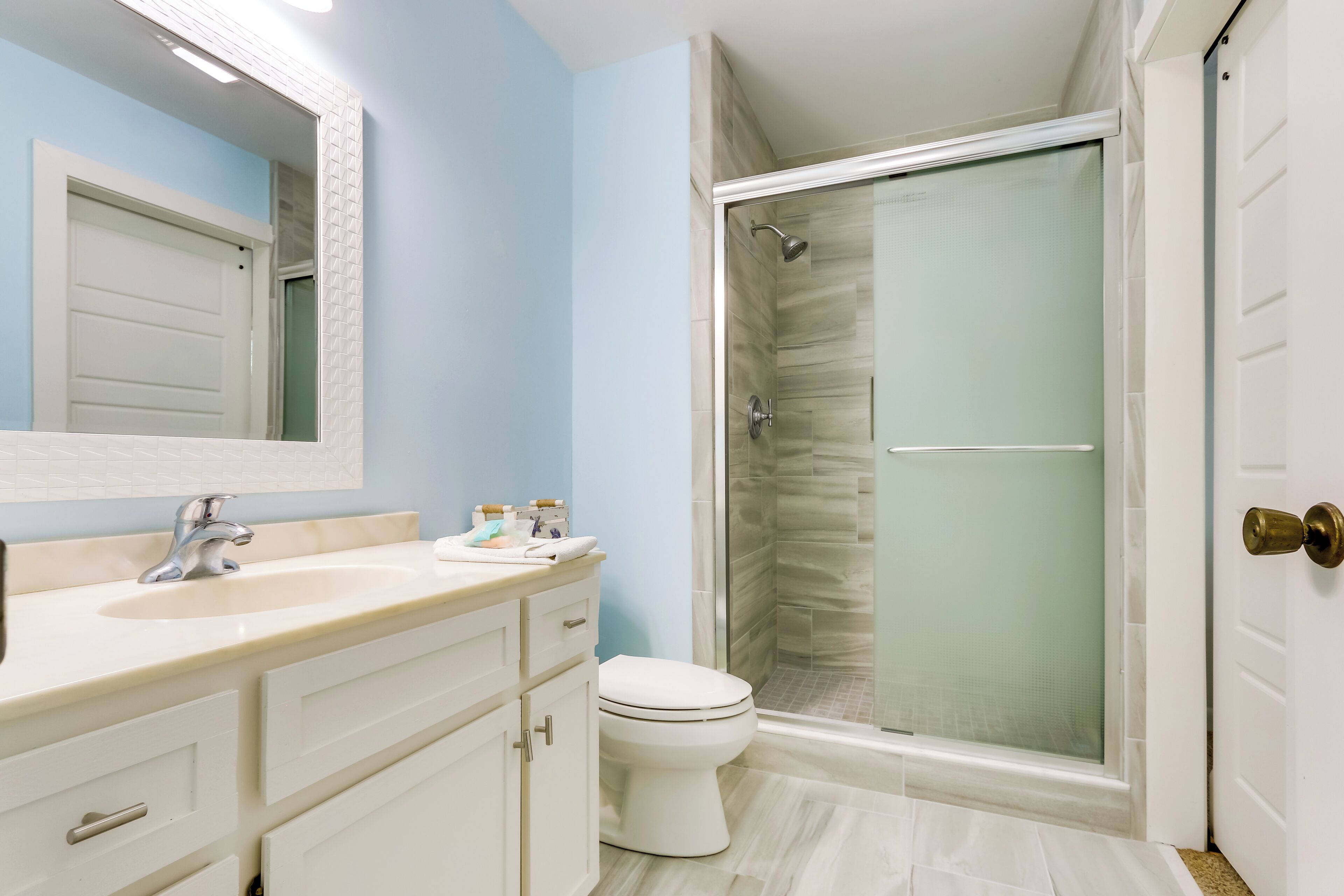 Une salle de bain avec un lavabo, un miroir et des toilettes blancs, une douche avec des portes en verre et une porte blanche.