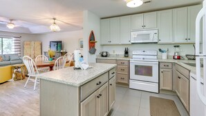 A modern kitchen with light-colored cabinets, a granite countertop, and stainless steel appliances. There is a dining area with a yellow sofa and white chairs.