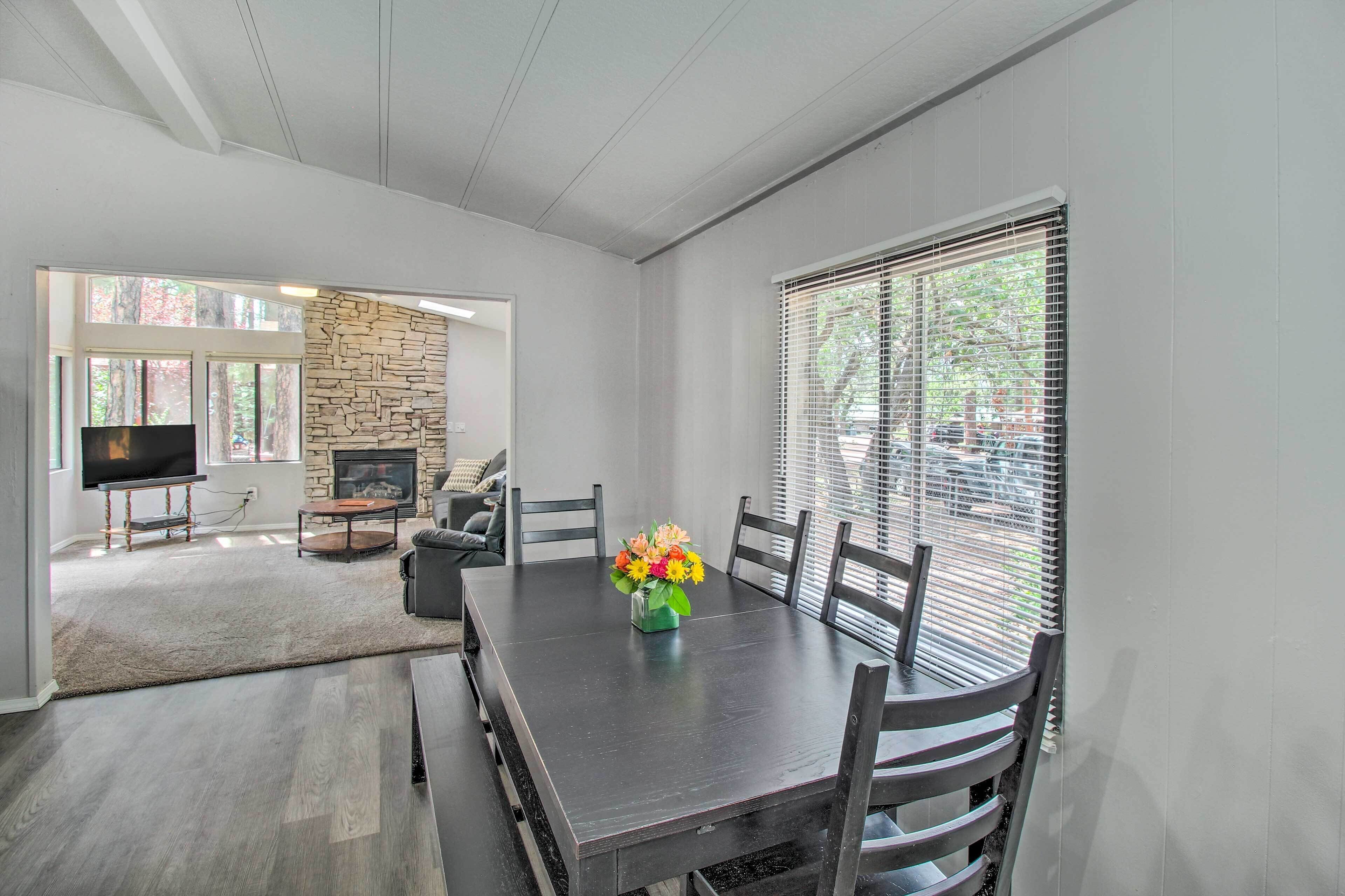 A modern dining area with a dark wooden table, six chairs, and a vase of flowers. A stone fireplace and a television are visible in the background.