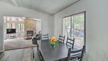 A modern dining area with a dark wooden table, six chairs, and a vase of flowers. A stone fireplace and a television are visible in the background.