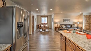 A kitchen with a stainless steel refrigerator, wooden dining table, and a living area with a sofa and lamp.