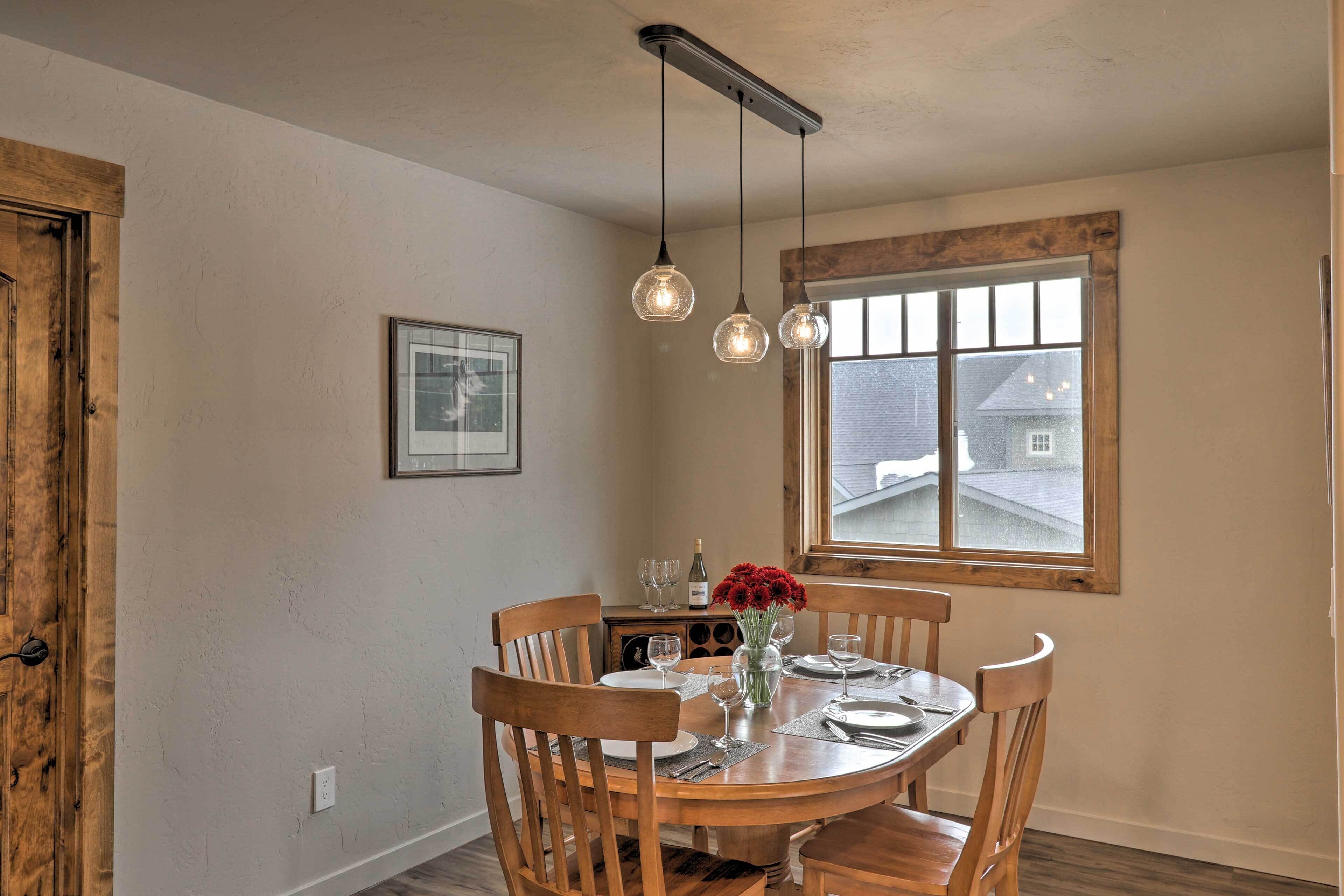 A dining area with a wooden table, chairs, hanging pendant lights, and a window with a view of a house.