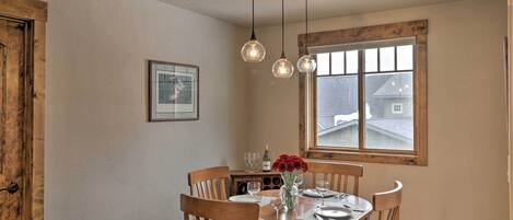 A dining area with a wooden table, chairs, hanging pendant lights, and a window with a view of a house.