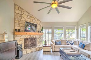 A living room with a stone fireplace, a ceiling fan, and a large window with blinds. - Horseshoe Bay Townhome w/ Patio & Outdoor Pool! (Horseshoe Bay)