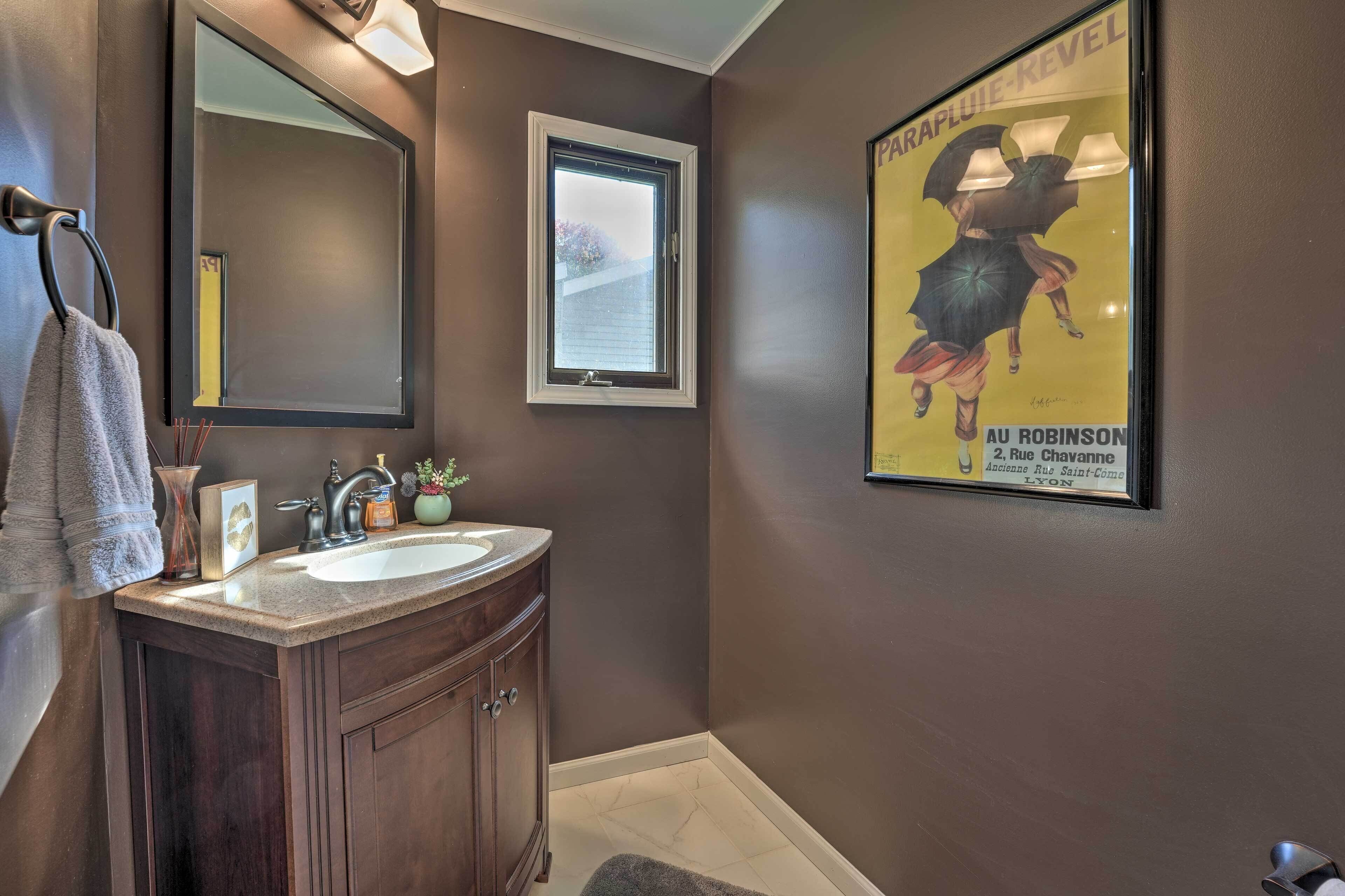 A bathroom with a dark brown wall, a large mirror, a window, a sink with a vanity, and a poster on the wall.
