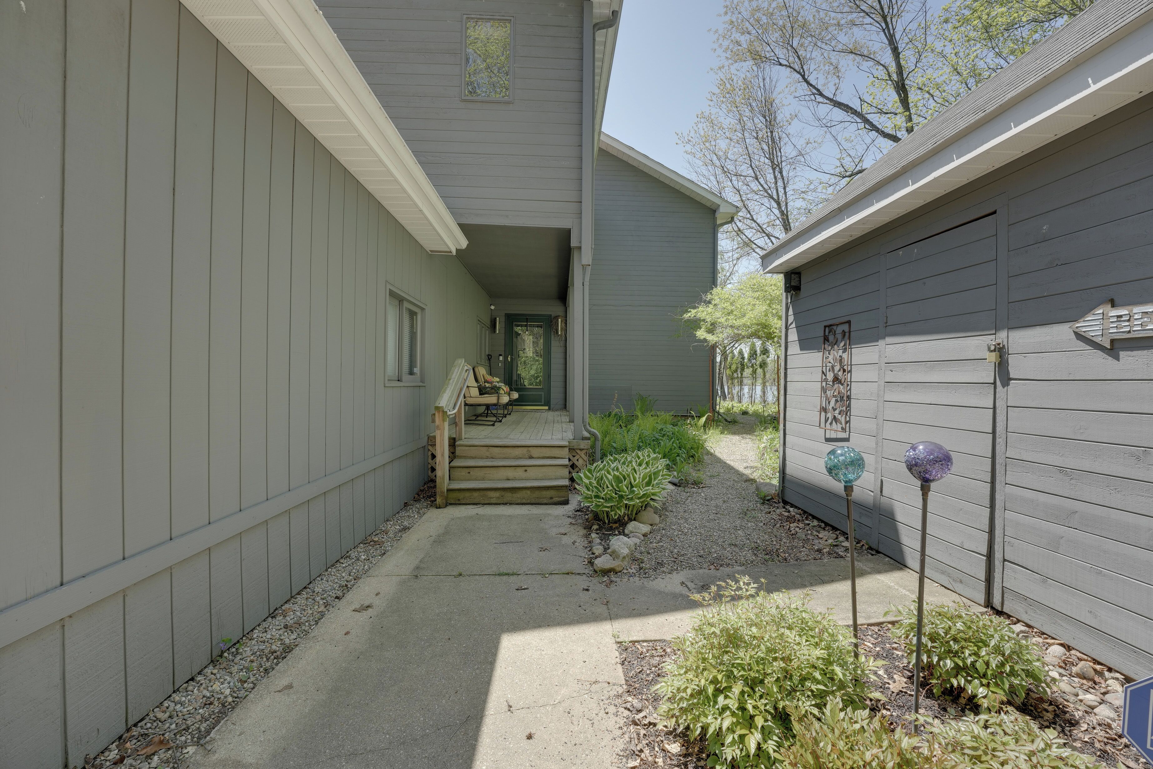 A narrow pathway leading to a wooden front door with steps, surrounded by gravel and small plants.