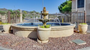 Una fuente de piedra circular con chorros de agua, rodeada de grava, con una cerca y un edificio al fondo.