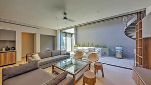 A modern living room with a glass coffee table, sofas, and wicker chairs. A kitchen area with a grill is visible in the background. - Upscale Cabo Home w/ Jacuzzi, Above Puerto Paraiso (Cabo San Lucas)