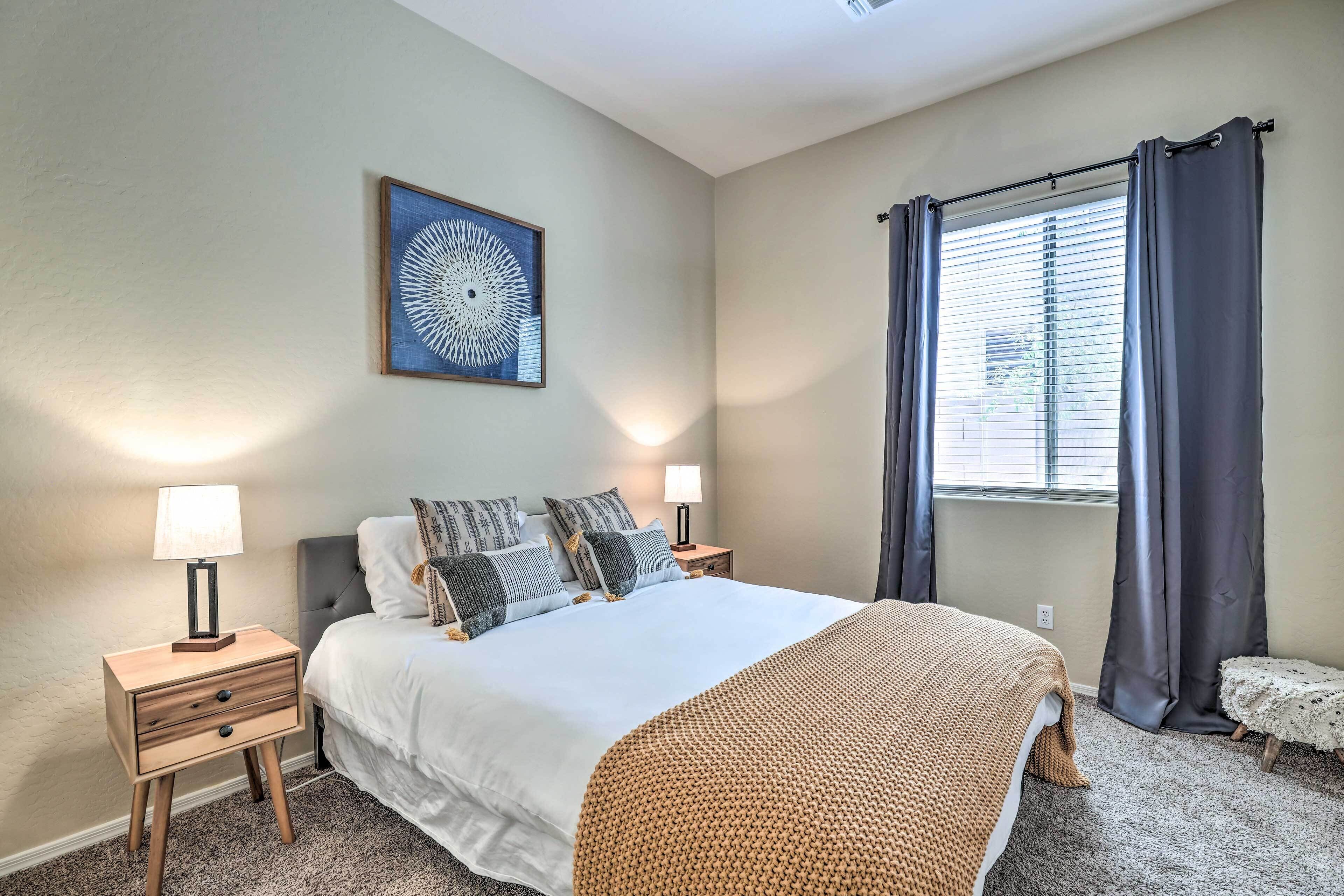 A bedroom with a bed, two bedside tables, a nightstand, a wall-mounted lamp, a window with curtains, and a wall art piece.