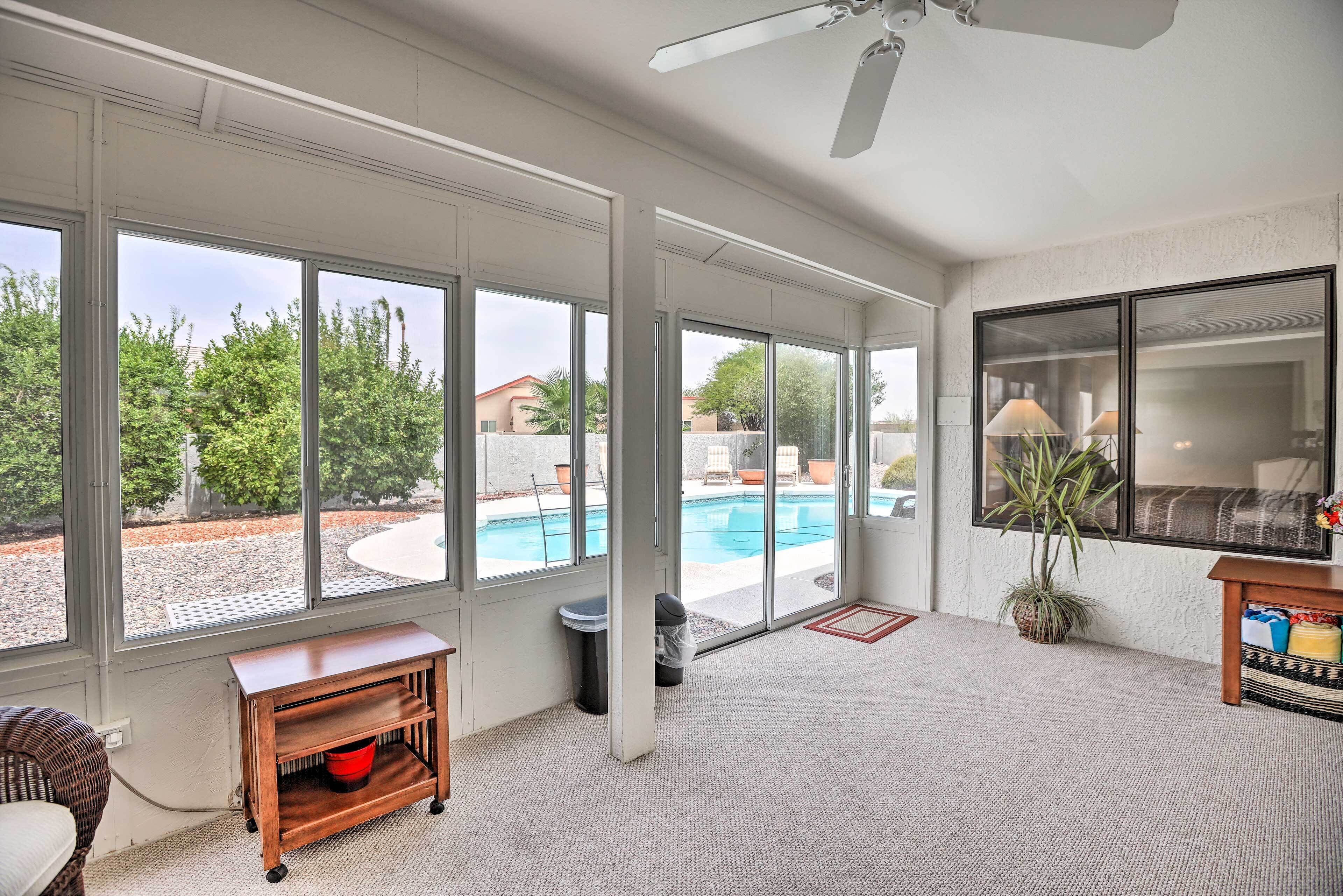 Amplia habitación con ventanas grandes, ventilador de techo, una pequeña mesa de madera y vista a una piscina exterior.