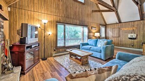 A cozy living room with wooden walls, a television, a blue sofa, and a coffee table. - Game Room & Fire Pit: Tree-lined Pocono Lake Cabin (Pocono Lake)