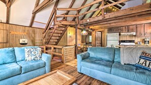 A rustic interior with wooden beams, a staircase, a kitchen, and two blue sofas. - Game Room & Fire Pit: Tree-lined Pocono Lake Cabin (Pocono Lake)
