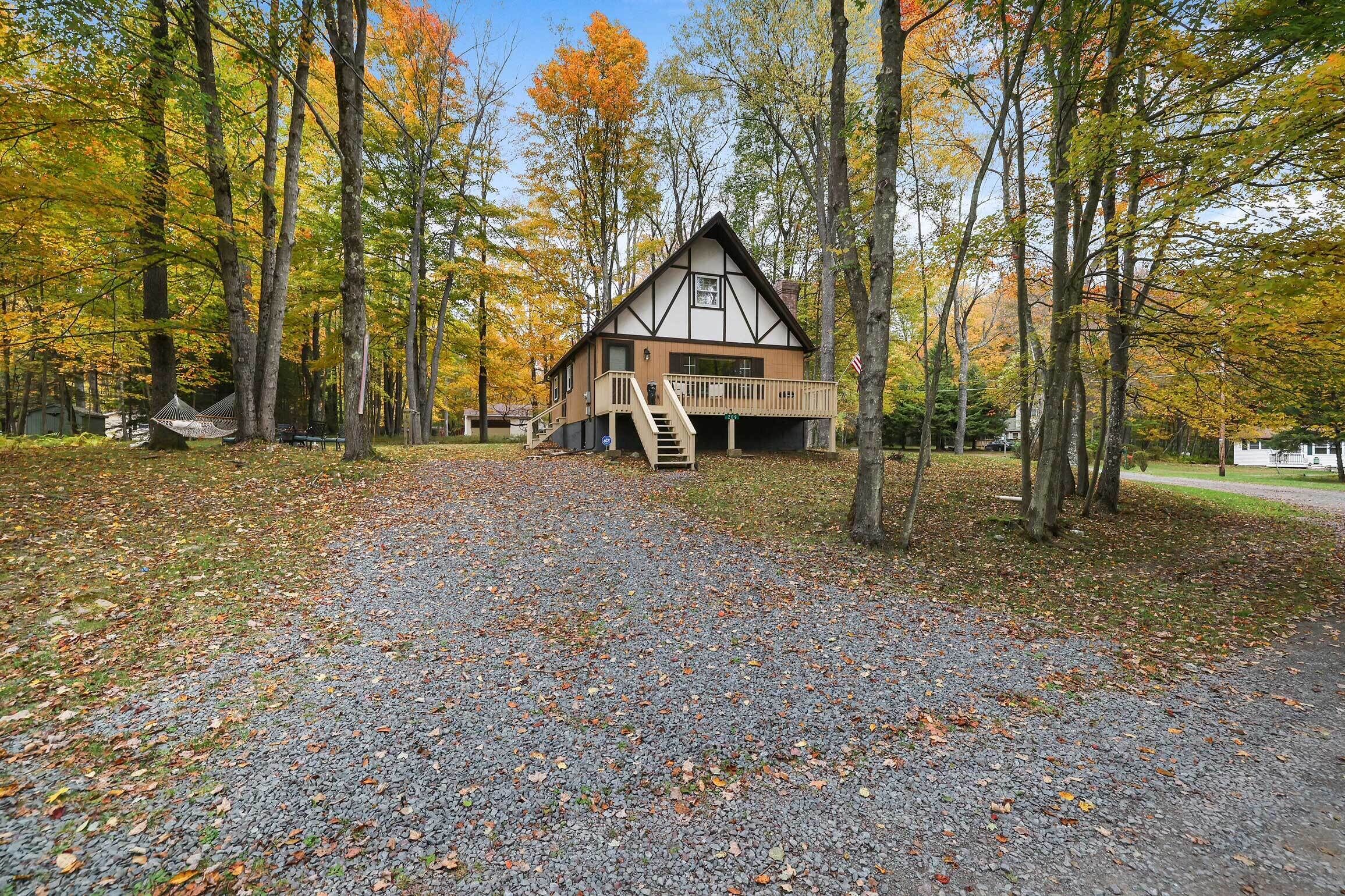 En hytte med træterrasse, trappe og grusvej omgivet af træer.