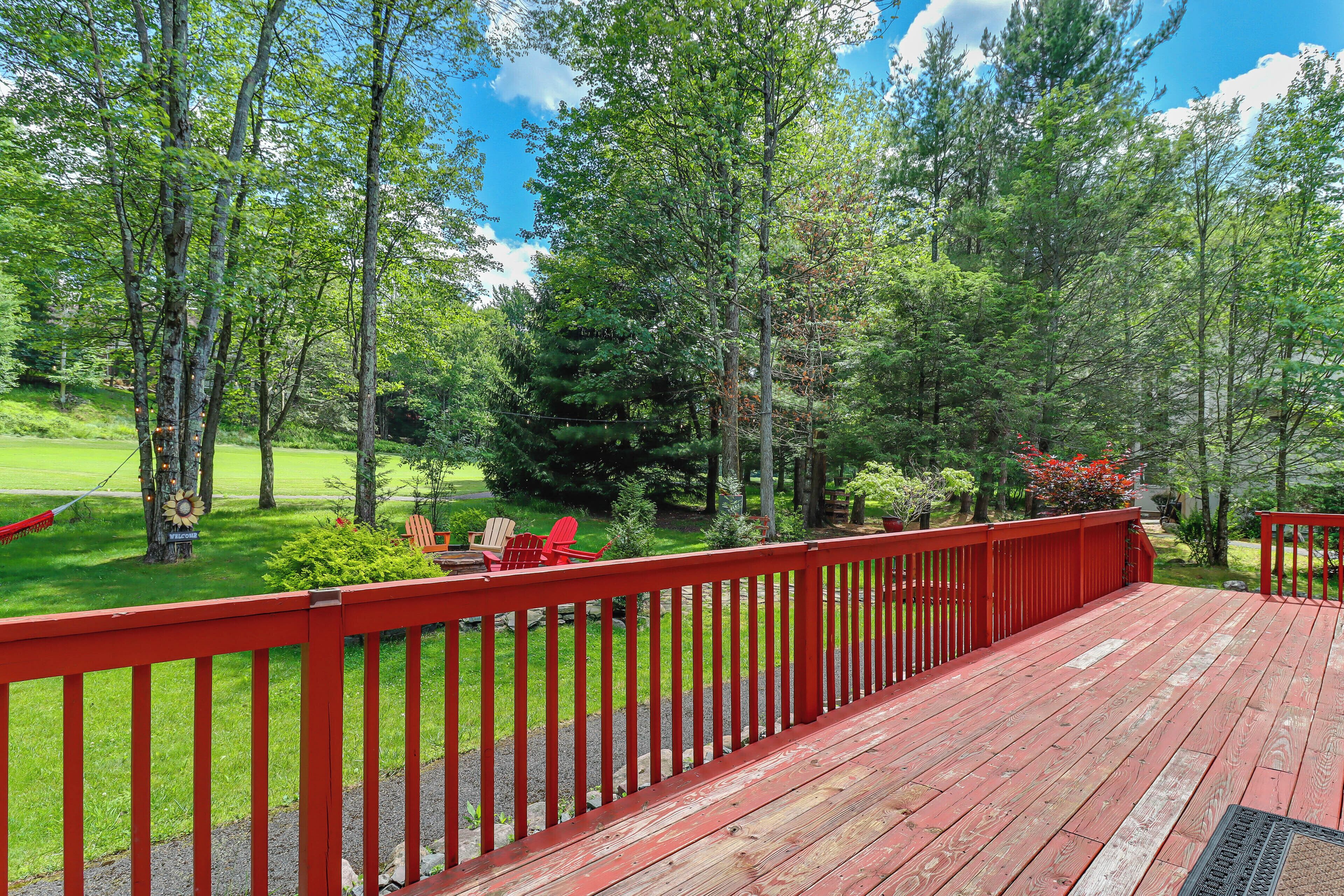 A wooden deck with a red railing, surrounded by trees and a grassy area with red chairs.
