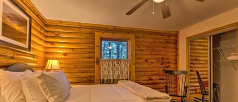A log cabin bedroom with a bed, a nightstand, a window with curtains, a ceiling fan, and a wooden wardrobe.