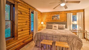 Quarto de cabana de toras com uma cama, um ventilador de teto e uma janela com cortinas.