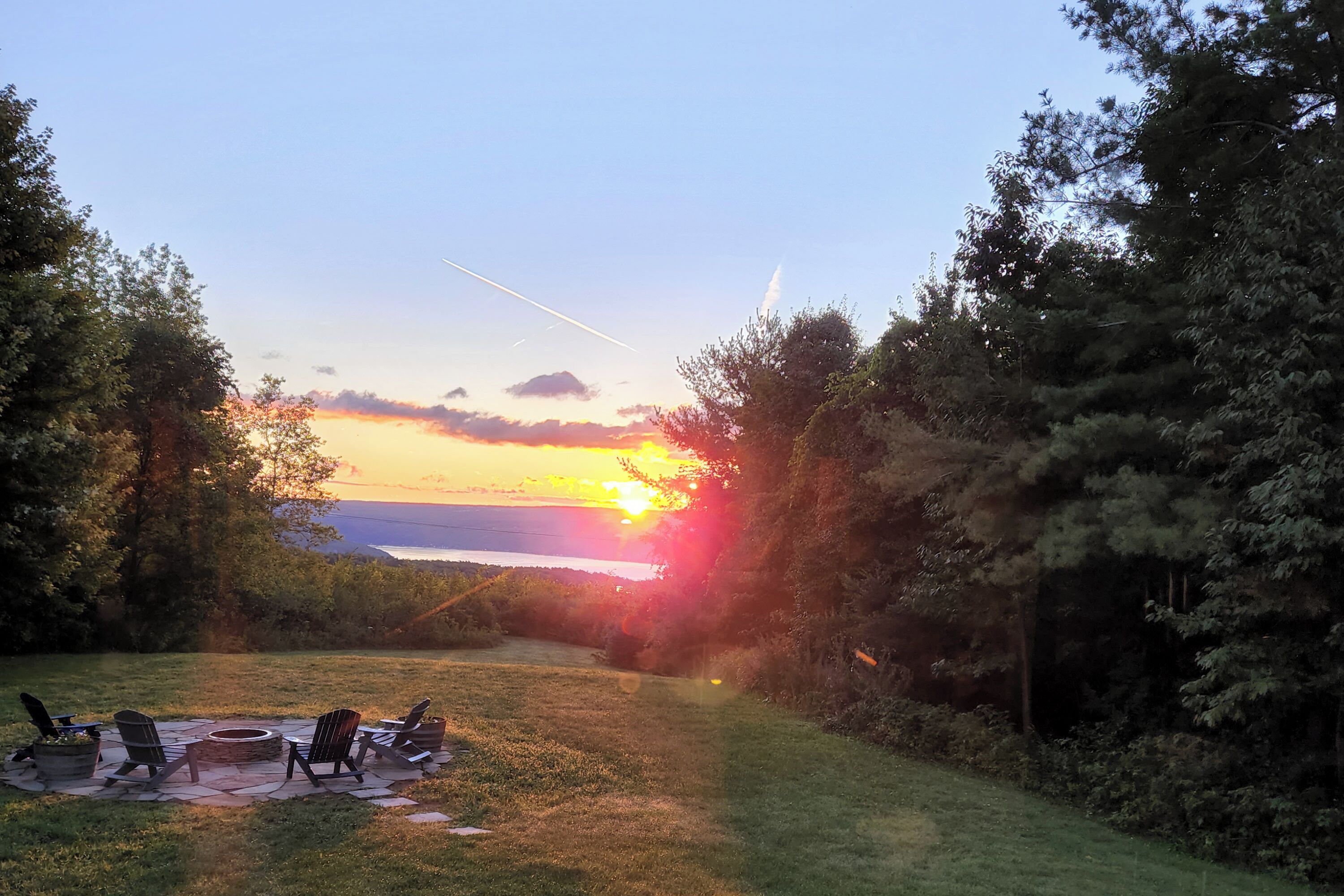A patio with chairs and a fire pit, surrounded by trees and a sunset view.
