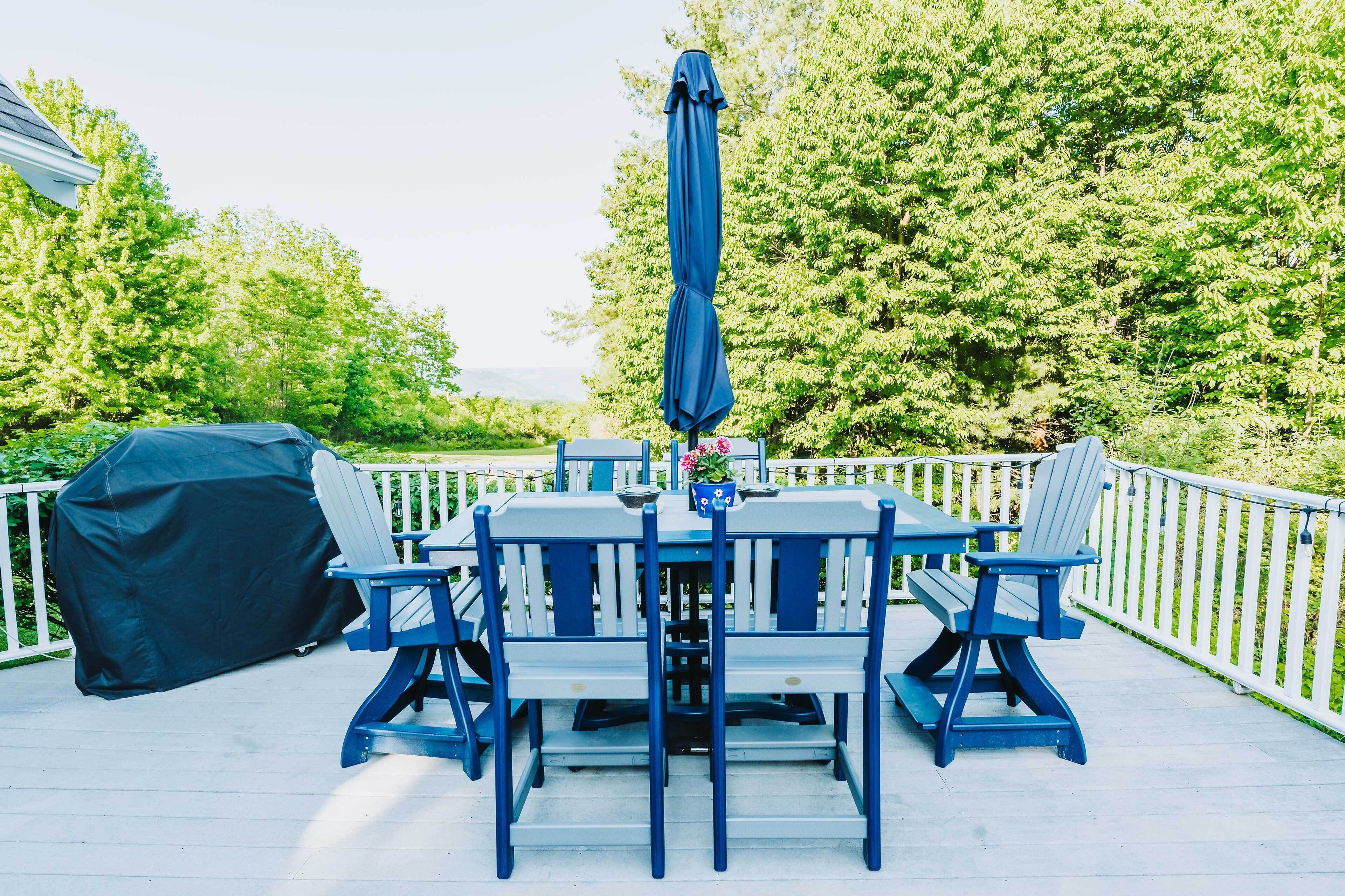 A covered outdoor deck with a table and chairs, surrounded by a white fence and lush greenery.