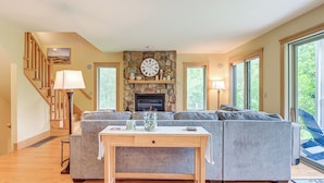 A living room with a stone fireplace, a gray sectional sofa, a wooden coffee table, and a staircase with wooden railings. - Finger Lakes Home w/ Private Pool & Lake Views! (Hammondsport)