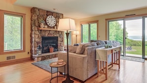 A cozy living room with a stone fireplace, a sofa, a coffee table, and large windows offering a view of the outdoors. - Finger Lakes Home w/ Private Pool & Lake Views! (Hammondsport)