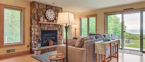 A cozy living room with a stone fireplace, a sofa, a coffee table, and large windows offering a view of the outdoors.