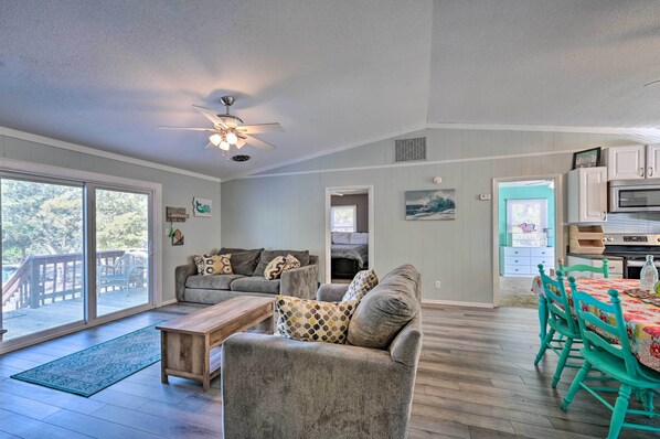 A spacious living room with a gray sofa, wooden coffee table, and teal chairs around a dining table. A ceiling fan and large windows provide natural light.