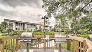 An outdoor deck with two barbecue grills, a wooden fence, and a view of a building in the background. - Walk to Beach: Resort-style Siesta Key Condo! (Siesta Key)