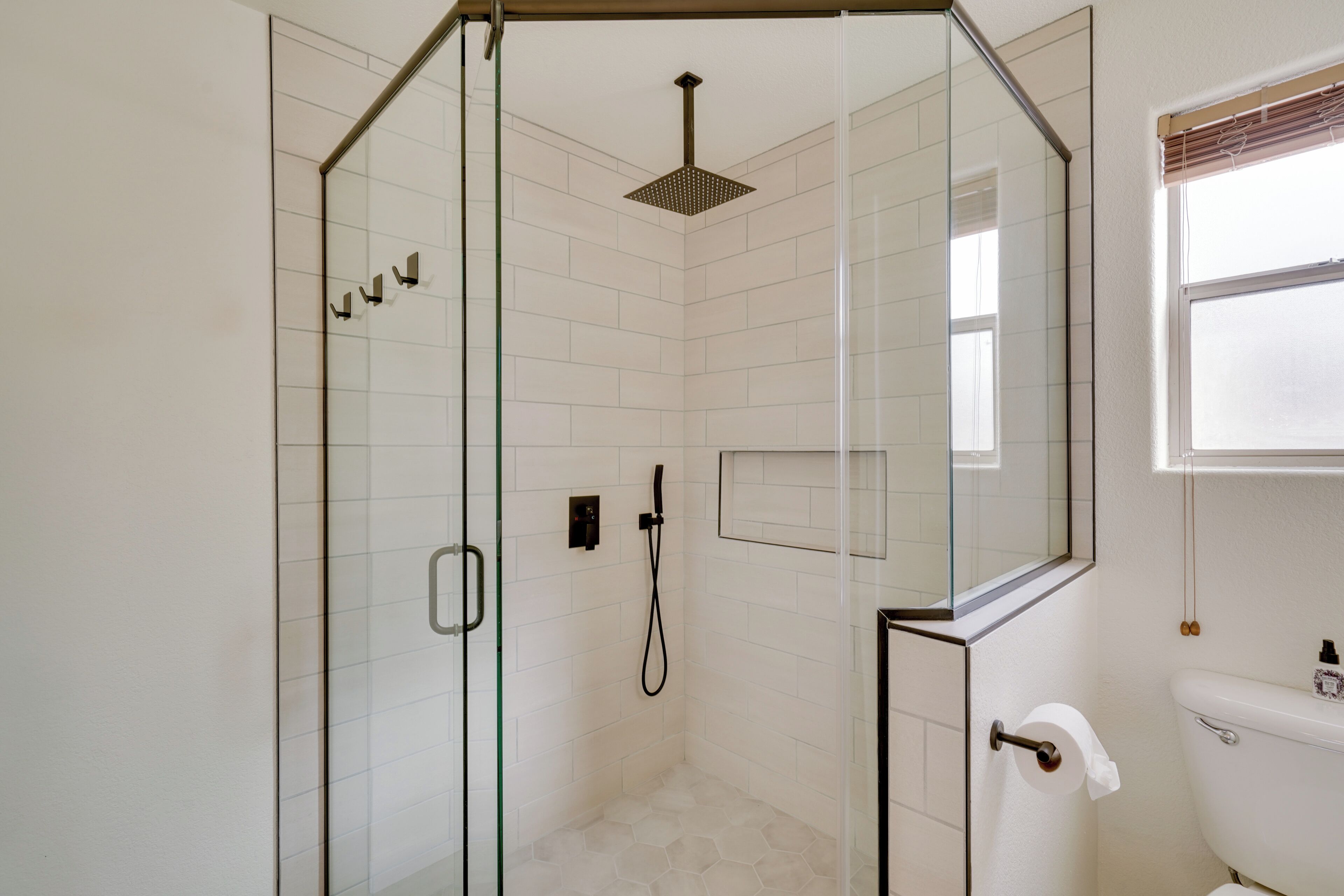 A modern bathroom with a glass-enclosed shower, white tiled walls, a toilet, and a window.