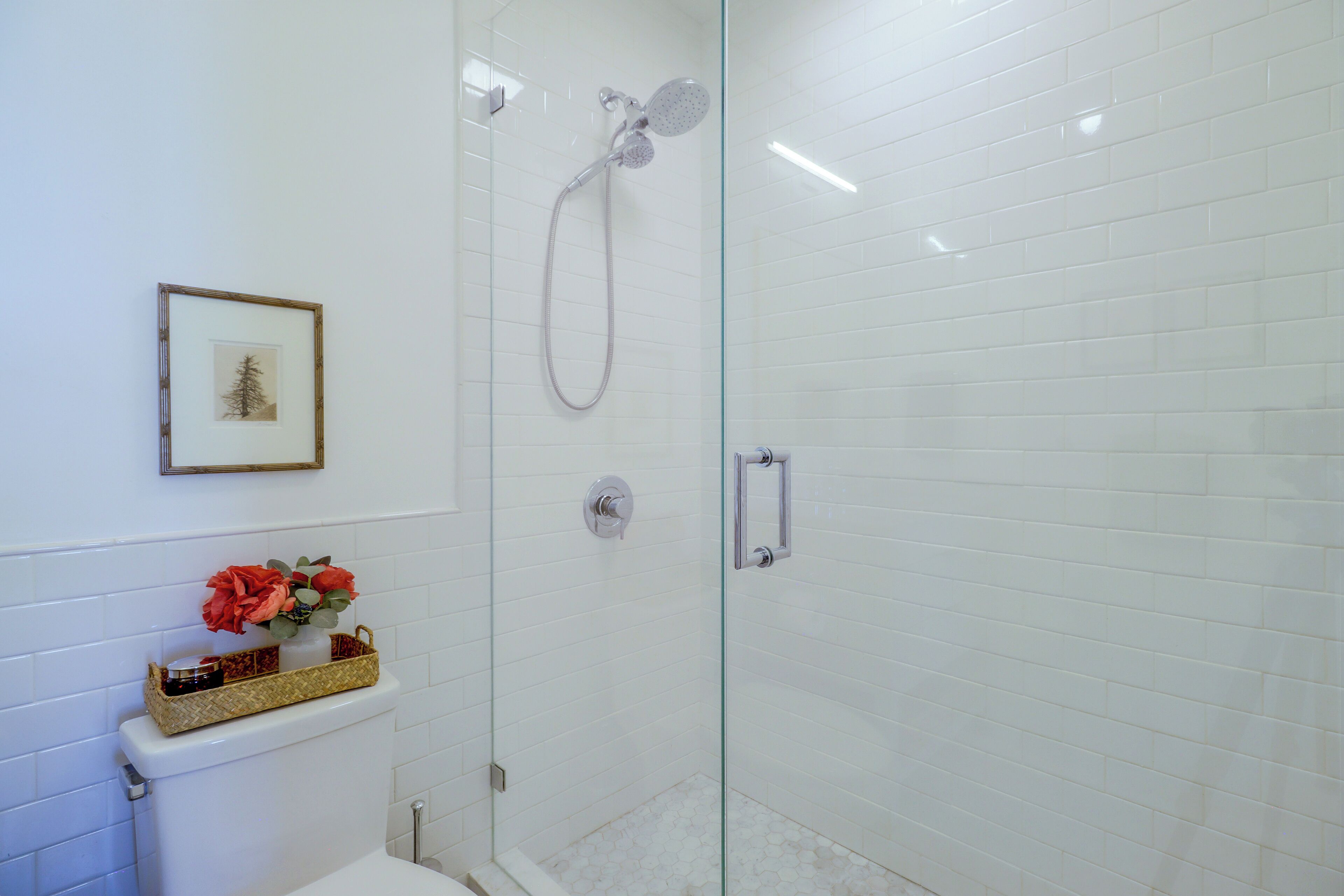 A bathroom with white tiled walls, a framed picture, a toilet with a basket of toiletries, and a glass-enclosed shower area.