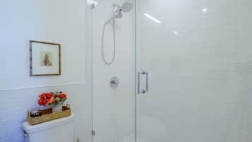 A bathroom with white tiled walls, a framed picture, a toilet with a basket of toiletries, and a glass-enclosed shower area.