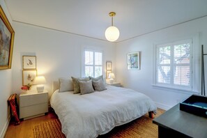 A bedroom with a large bed, two bedside tables with lamps, a window with blinds, and artwork on the walls. - Charming Midtown Brick Home: Walk to Truckee River (Reno)