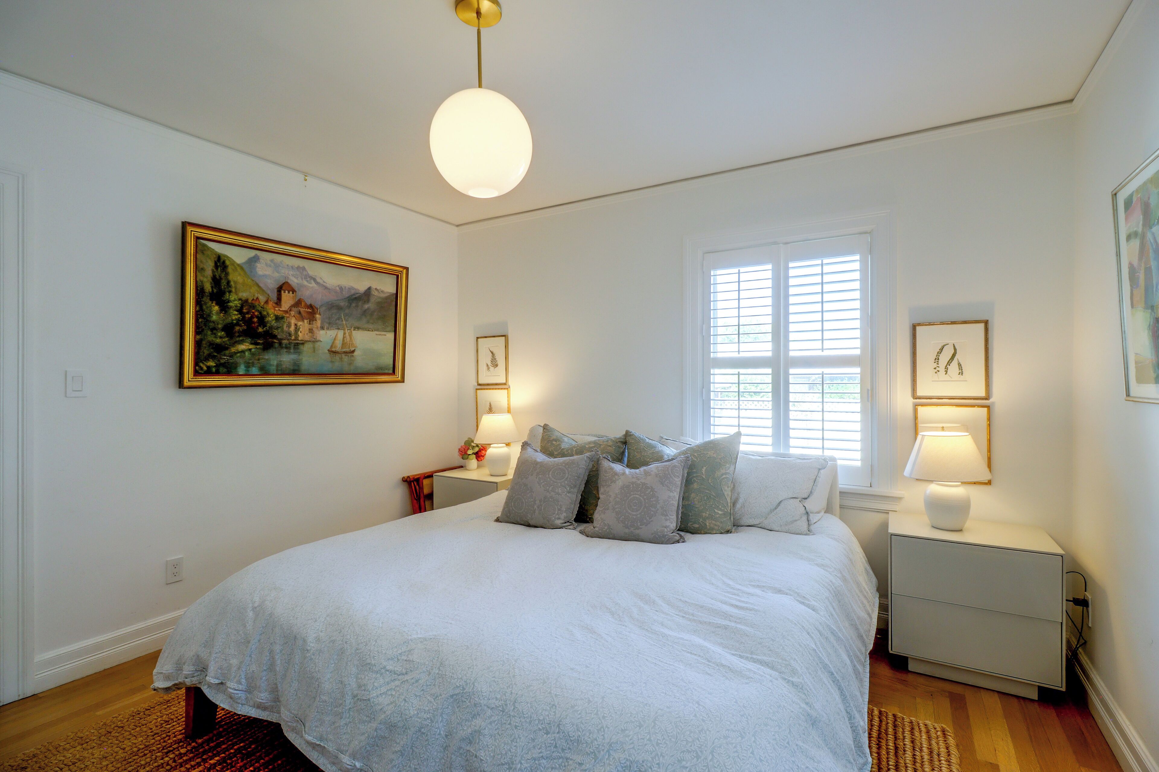 A bedroom with a large bed, a painting, a window with blinds, and two bedside lamps.