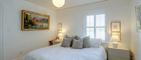 A bedroom with a large bed, a painting, a window with blinds, and two bedside lamps.