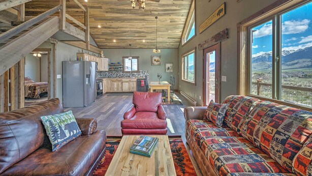 A spacious living area with a high ceiling, a red leather sofa, a patterned couch, a wooden coffee table, and a view of mountains through large windows.
