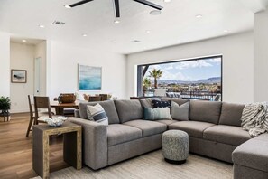 A modern living room with a large sectional sofa, a dining table, and a view of a pool and mountains through large windows.