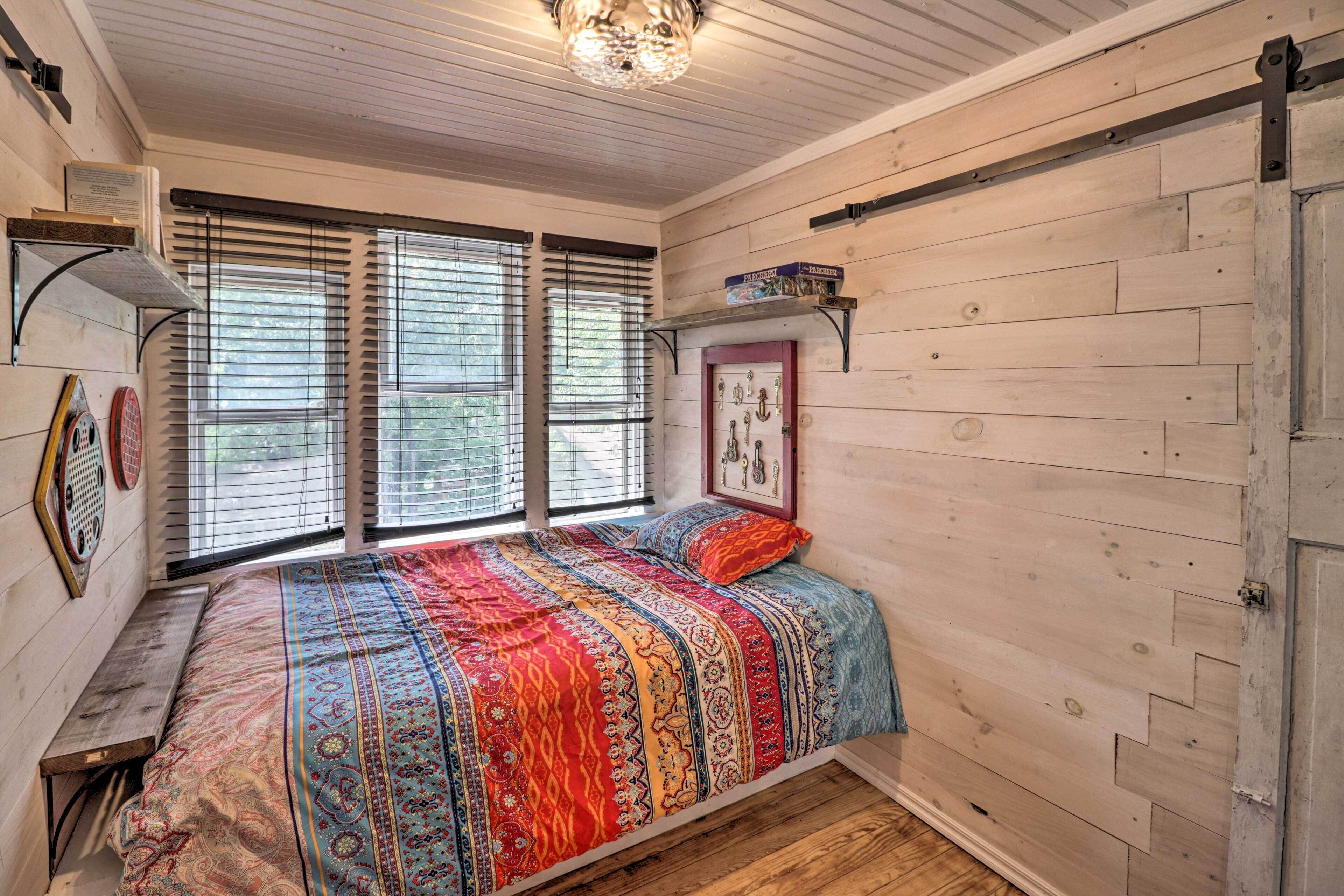 A bedroom with a wooden floor, a bed with a colorful blanket, a shelf with books, and a window with blinds.